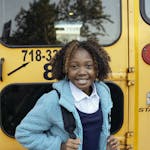 Cheerful African American girl with curly hair standing with backpack near yellow school bus and looking at camera with smile
